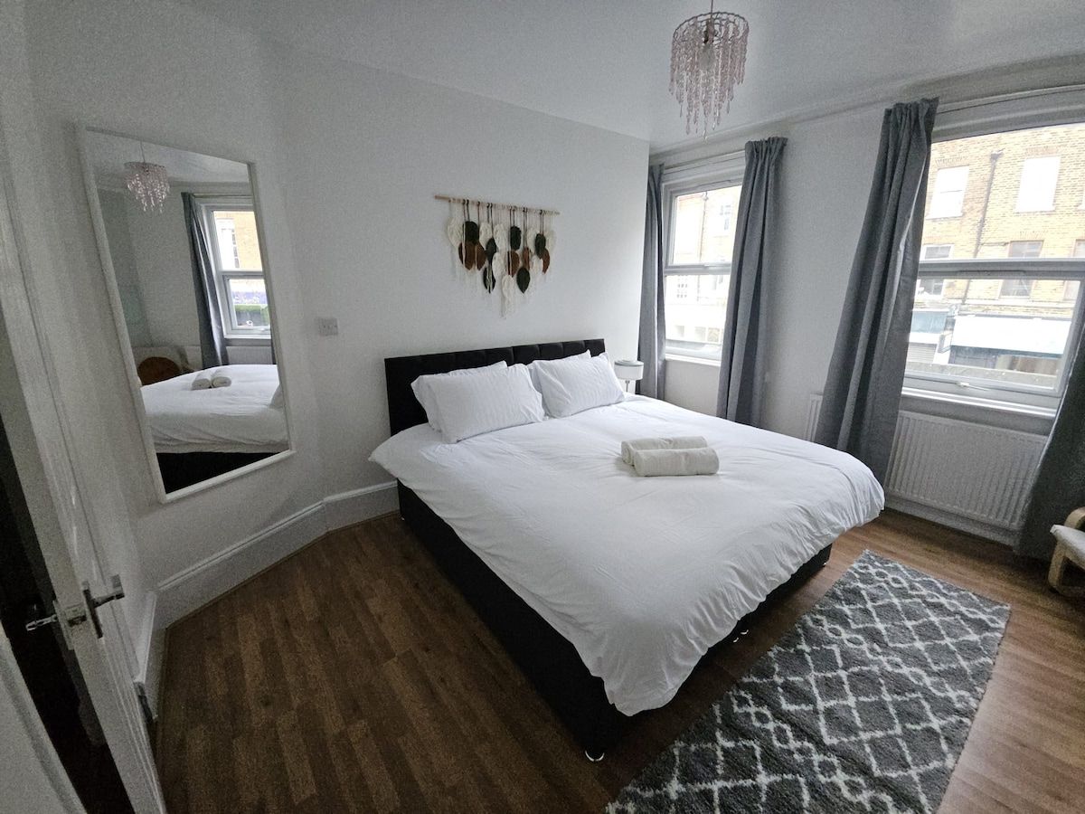 A bedroom with a large bed, a mirror, a wall-mounted decoration, and a view of the outside through a window.