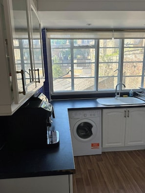 Une cuisine compacte équipée d’un lave-linge, d’une machine à café et d’une fenêtre donnant sur des immeubles.