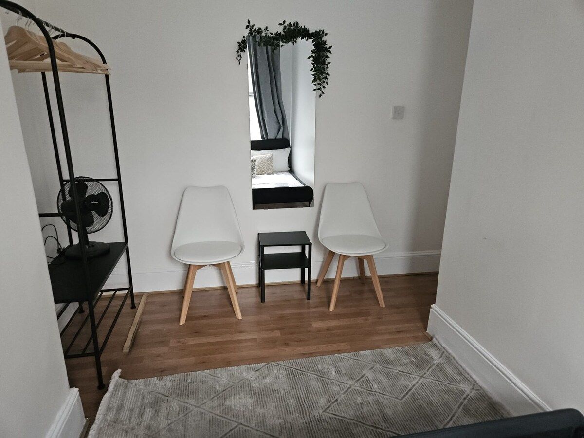 A small room with a bed, two white chairs, a black fan, a small table, and a window with curtains.