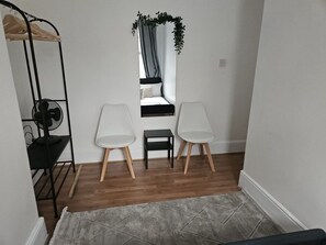 A small room with a bed, two white chairs, a black fan, a small table, and a window with curtains.