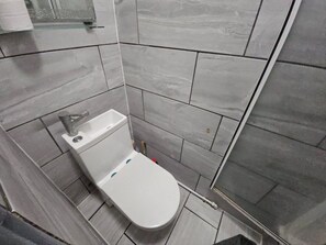 A bathroom with a white toilet, a glass shower enclosure, and grey tiled walls. - Central Location:2BR Flat Near Angel & City Centre (Greater London)