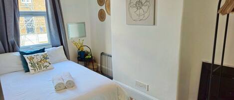 A neatly made bed with a white duvet and two rolled towels, a bedside table with a lamp, a radiator, and a wall-mounted artwork.