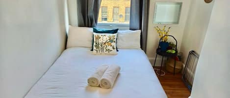 A neatly made bed with rolled towels, a window with curtains, a chair with a plant, and a radiator.