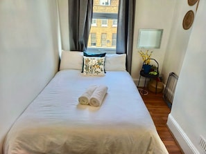 A neatly made bed with rolled towels, a window with curtains, a chair with a plant, and a radiator. - Central Location:2BR Flat Near Angel & City Centre (Greater London)