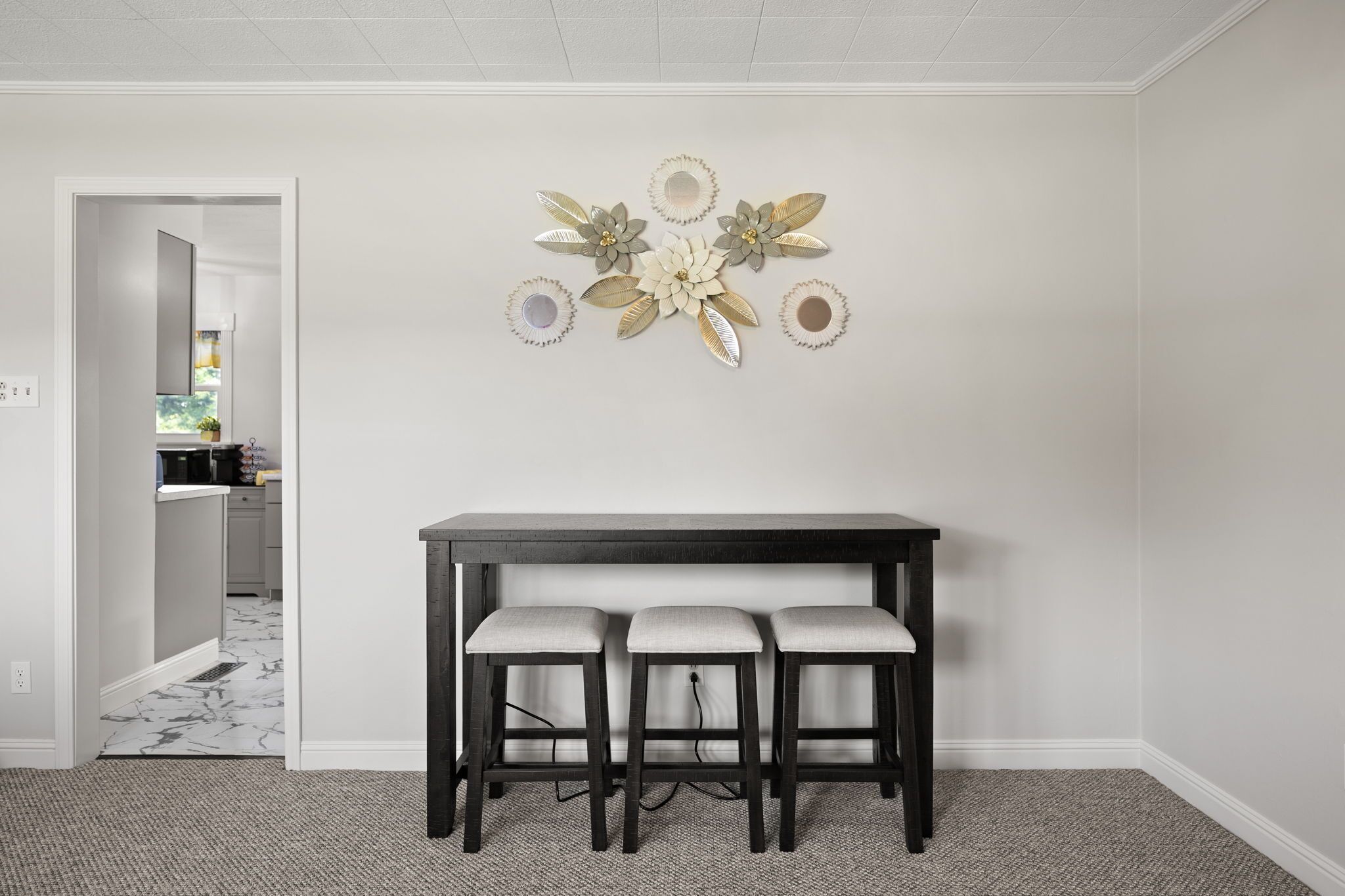 A dining area with a dark wooden table and three matching chairs, a decorative wall piece above, and a hallway leading to a kitchen.
