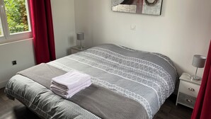 Un llit ben arrenjat amb un estenat de disseny, una tauleta de nit amb una làmpada, i una finestra amb cortines.