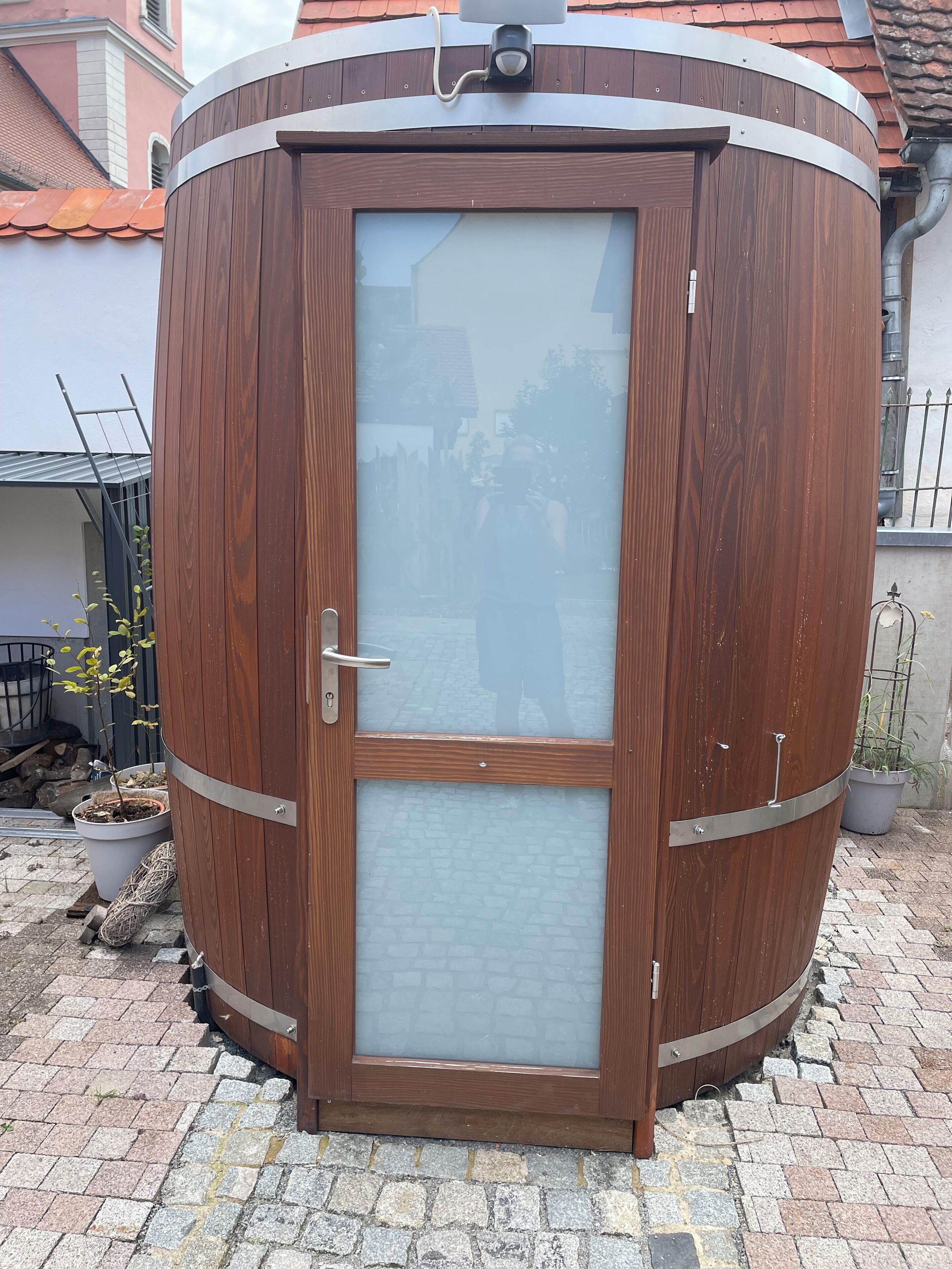 Una struttura cilindrica in legno con una porta in vetro, maniglie in metallo e un anello metallico nella parte superiore.