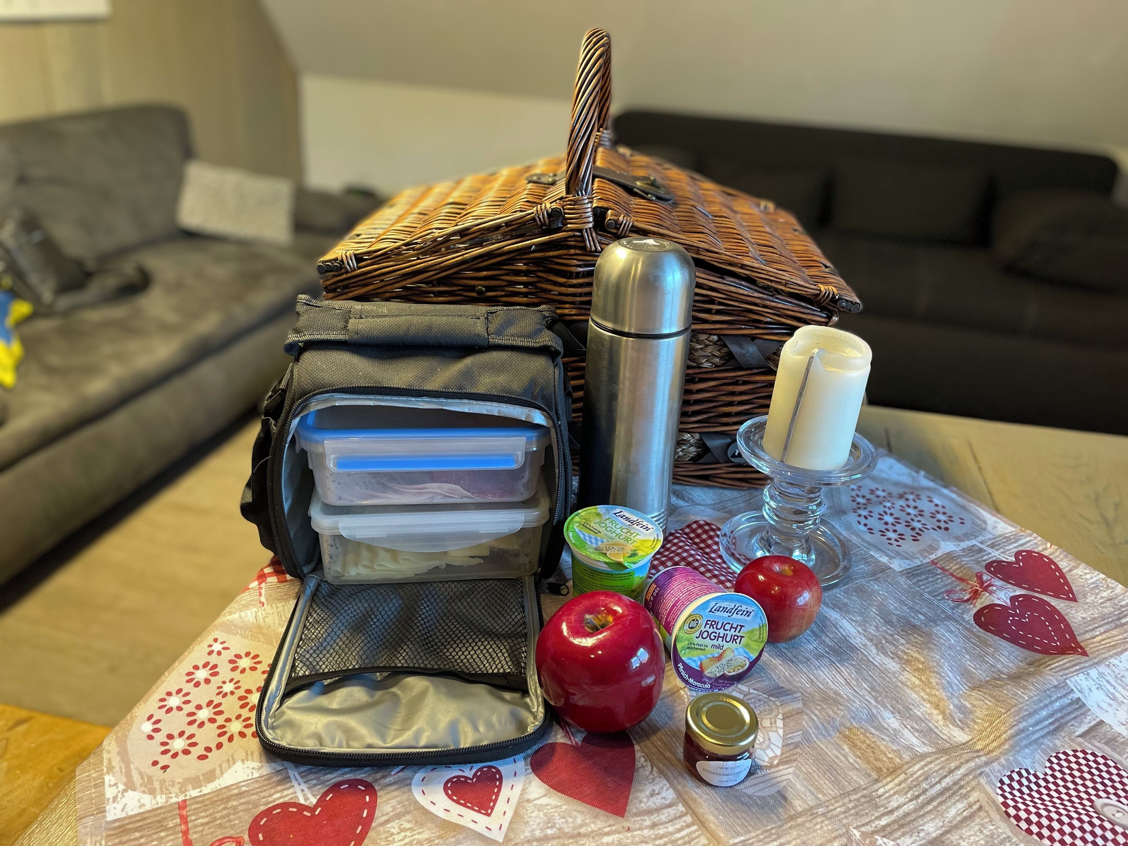 A wicker basket with a thermos, a candle, apples, and a jar of jam on a table with a patterned tablecloth.