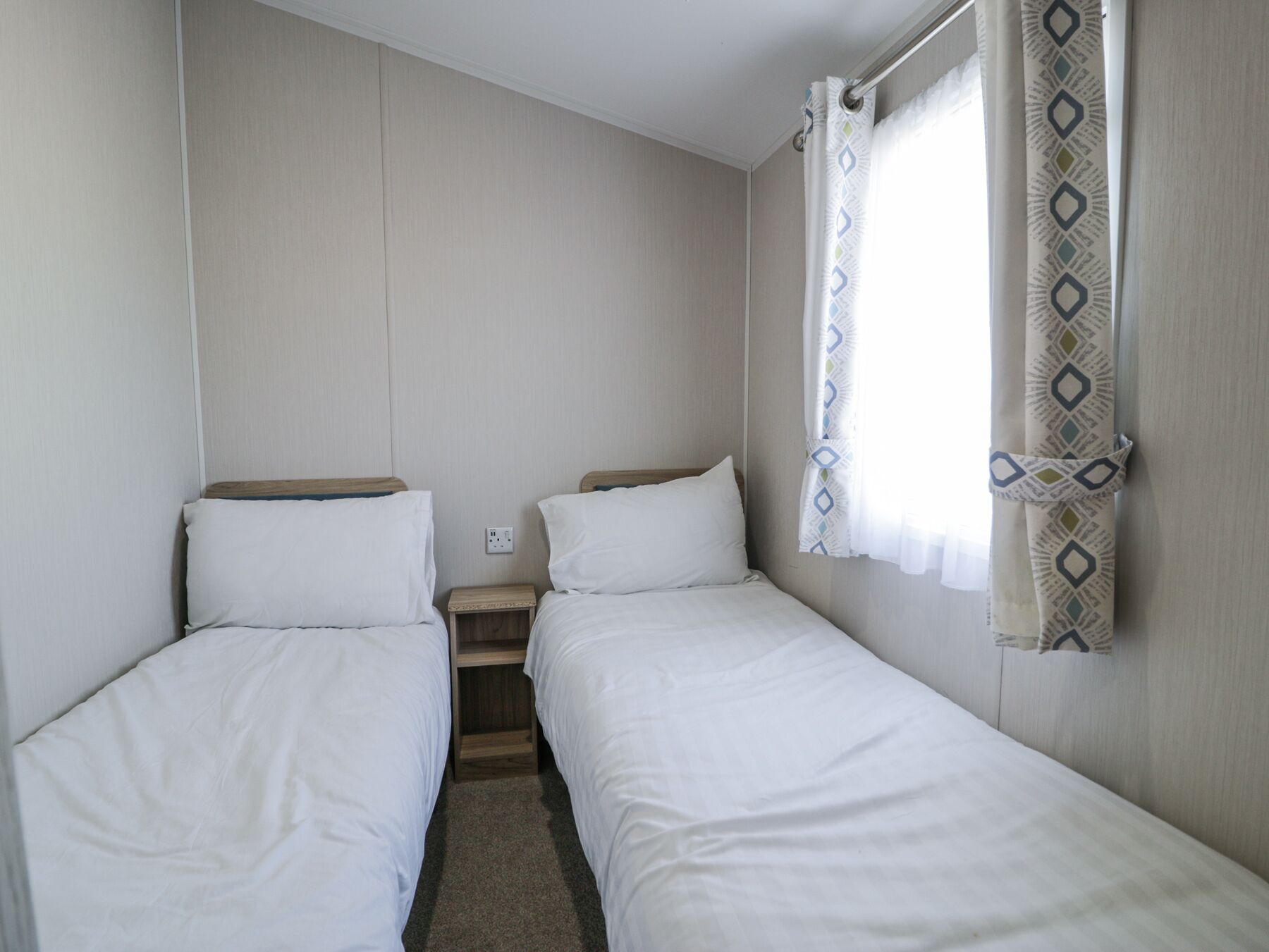 A small, single-room accommodation with two single beds, a small wooden nightstand, and a power outlet on the wall.