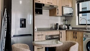A compact kitchen with a built-in refrigerator, microwave, and washing machine. There are two upholstered chairs around a small round table.