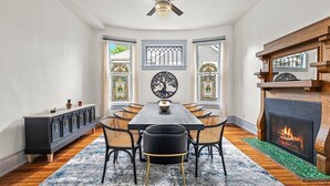 A dining room with a long table, six chairs, a chandelier, stained glass windows, and a fireplace.