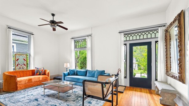 A spacious living room with a blue sofa, an orange sofa, a glass coffee table, and a wicker chair. There is a ceiling fan, a large window with a view of greenery, and a decorative mirror on the wall.