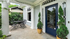 A covered porch with a dining table and chairs, a blue door, and two potted evergreen trees.