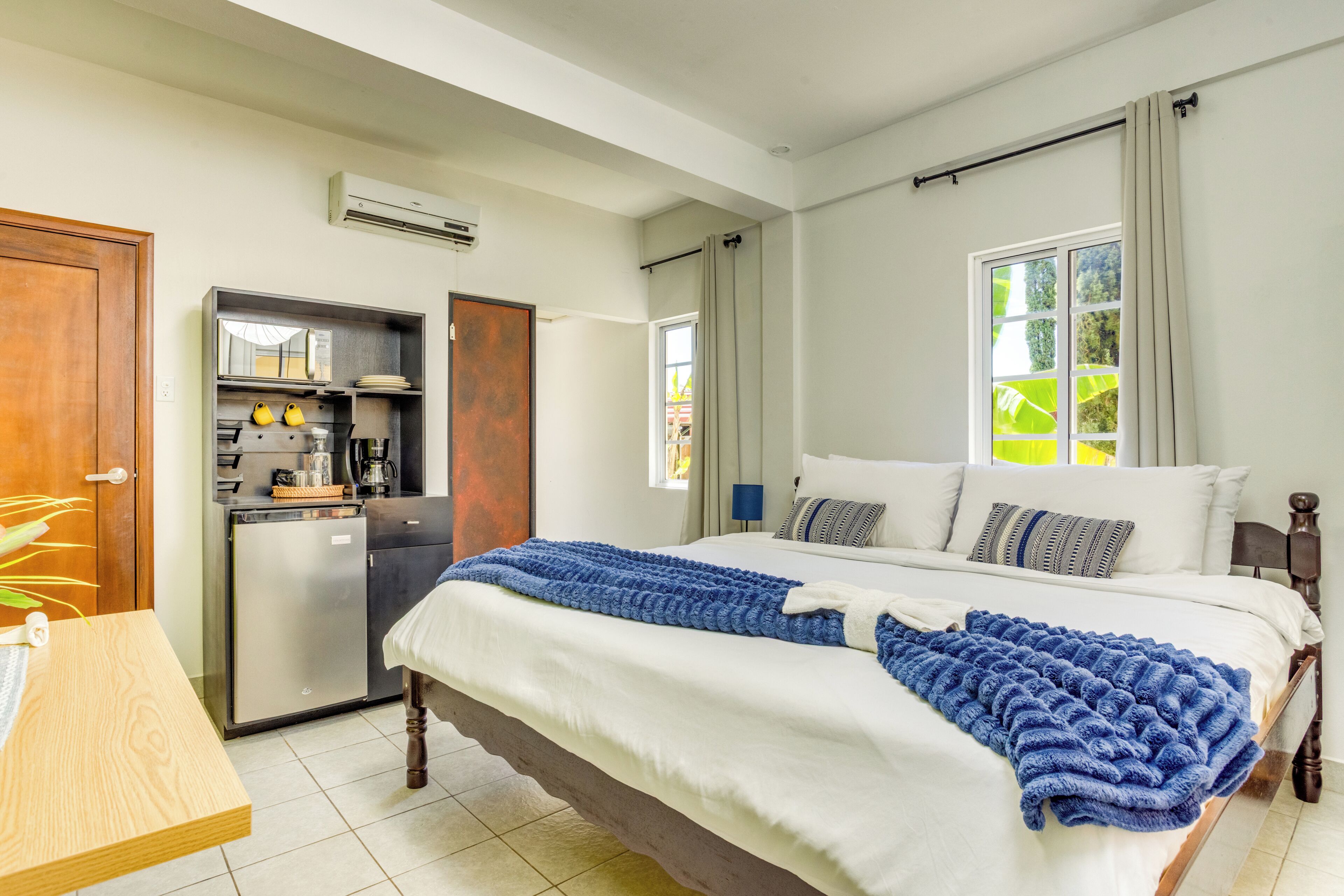 A bedroom with a bed, a desk, a kitchenette, and a window.