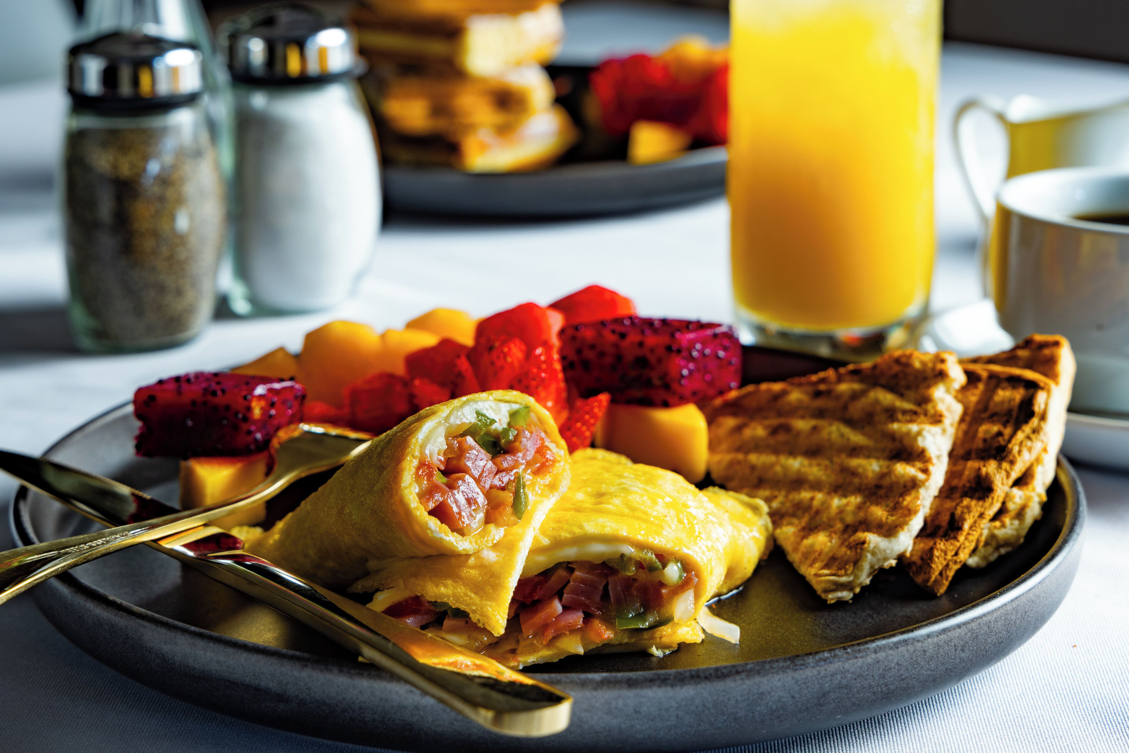 Prato de café da manhã com sanduíche, frutas e um copo de suco de laranja.