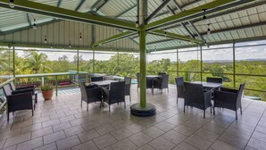 Een overdekt terras met tafels en stoelen, een potplant en uitzicht op groen.