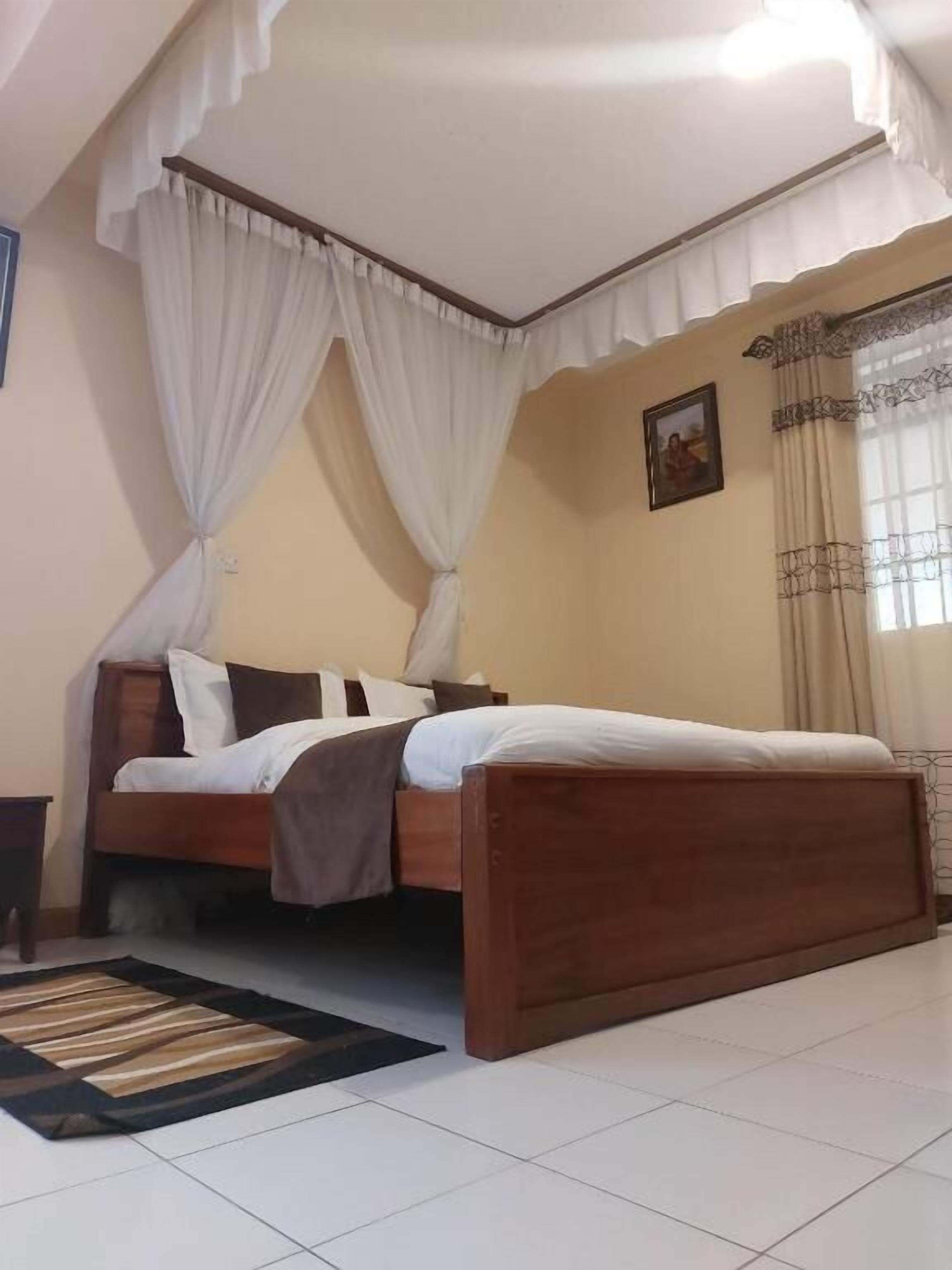 A bedroom with a wooden bed, white curtains, a picture on the wall, and a window with patterned curtains.