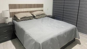 Ein ordentlich bezogenes Bett mit einer grauen Tagesdecke, einer Nachttischlampe und einem dunklen Nachttisch mit Schubladen.
