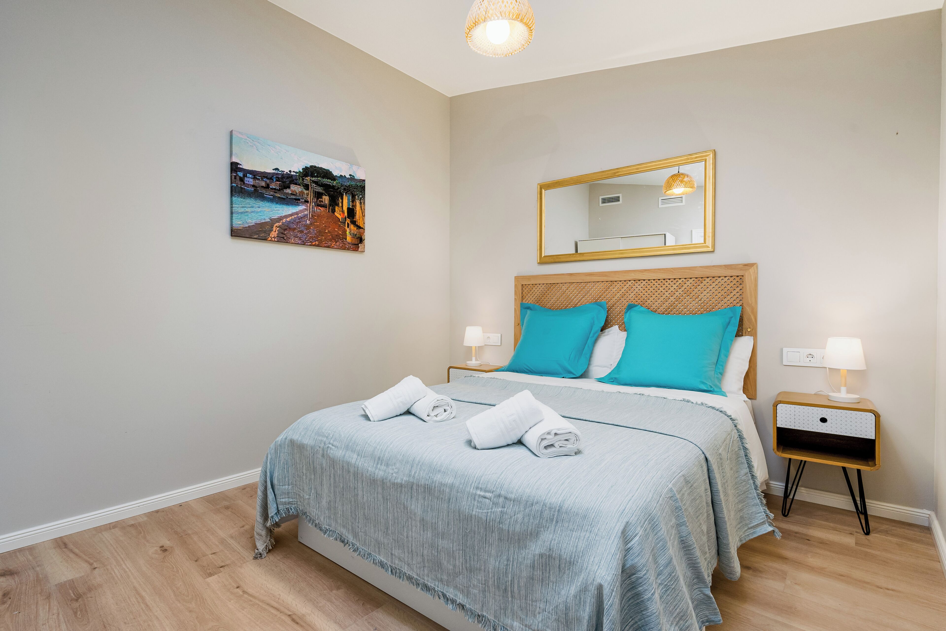 A neatly made bed with a light blue bedspread, two turquoise pillows, and two rolled towels on the bed. There is a small bedside table with a lamp, a wall-mounted mirror, and a painting of a coastal scene.