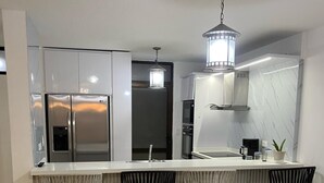Una cocina moderna con un refrigerador de acero inoxidable, horno empotrado, microondas y estufa a gas. Hay tres taburetes negros y una lámpara colgante.