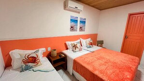 A room with two beds, each with a unique sea-themed pillow. There is a small table with a basket and a remote control, and an air conditioning unit mounted on the wall.