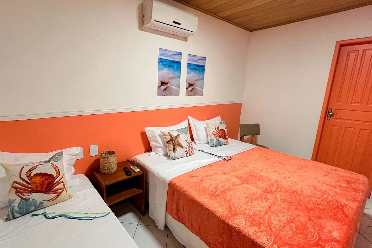 Quarto com duas camas, cada uma com almofada com tema náutico. Há uma pequena mesa com uma cesta e um controle remoto, e um ar condicionado fixado na parede.