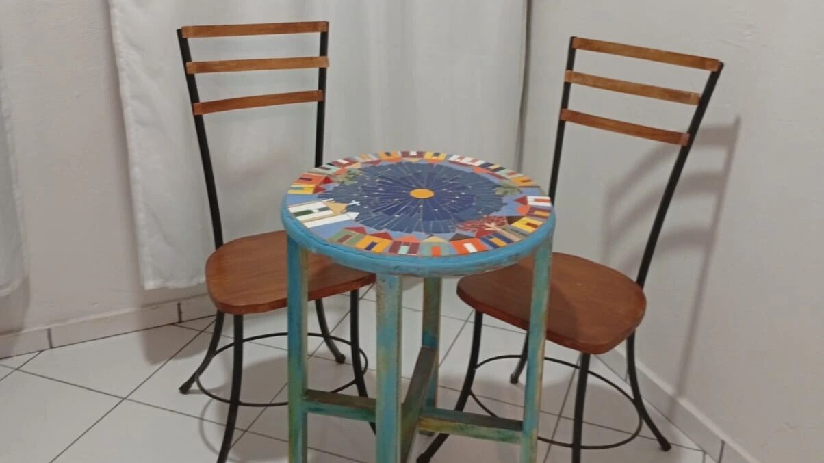 Una mesa redonda con una tapa de mosaico de colores, acompañada por dos sillas de madera con estructura metálica.