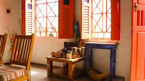 A room with a wooden chair, a small table with various items, and a window with orange shutters.