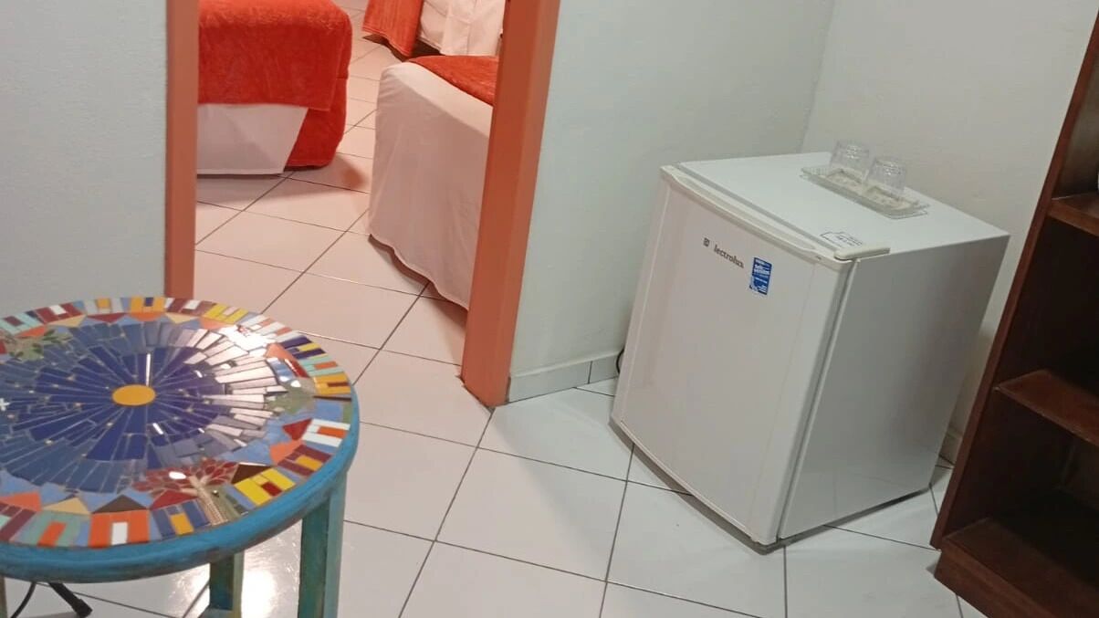 Una habitación con piso de baldosas, un refrigerador blanco pequeño, una mesa de mosaico colorido y una cama con ropa de cama naranja.