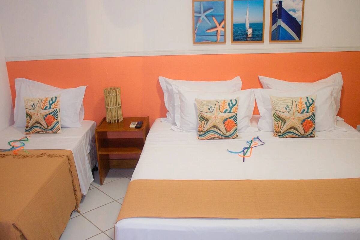 Quarto de hotel com duas camas, cada uma adornada com almofadas decorativas combinando, uma mesa de cabeceira com abajur e uma pequena mesa.