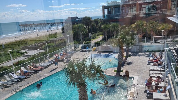 Zona de piscina con sillas de descanso, una palmera y vistas a la playa y al muelle.