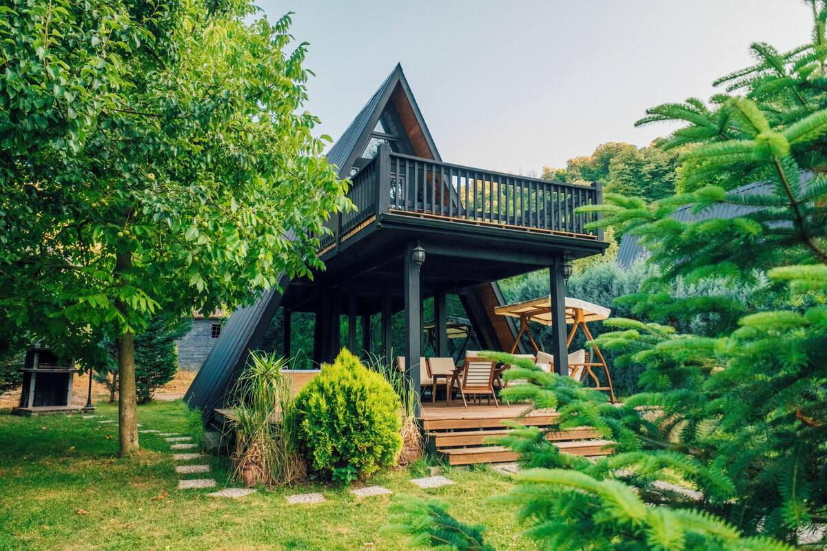 A modern house with a wooden deck, outdoor seating, and a large tree in the foreground.