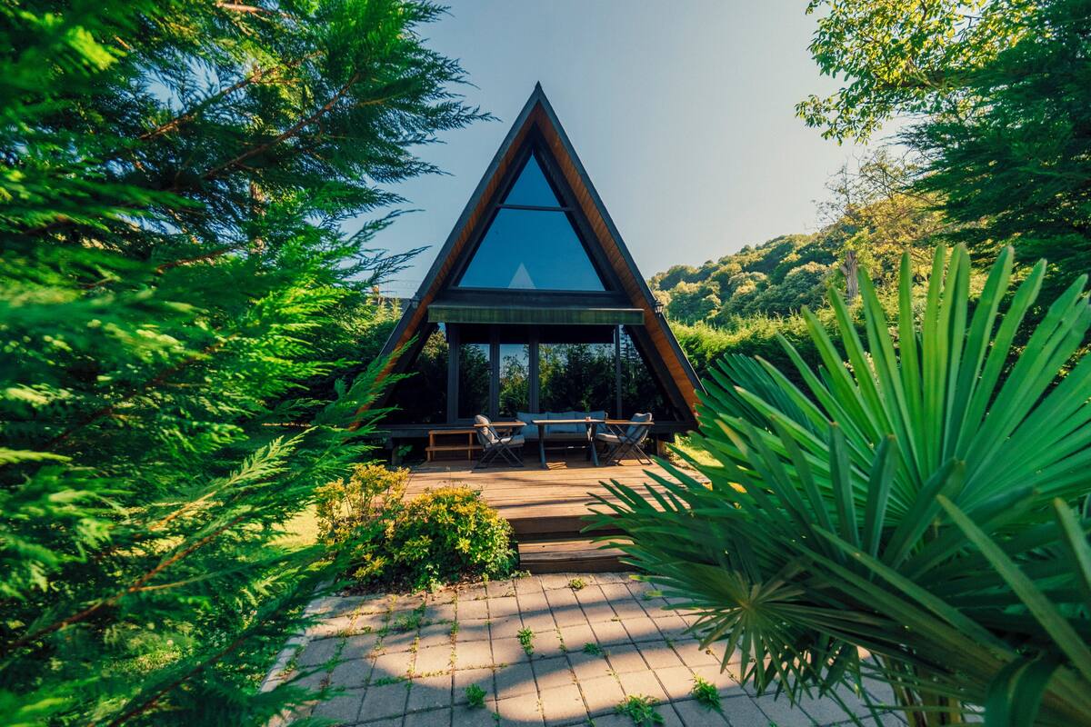 A triangular wooden structure with a glass front, surrounded by lush greenery.