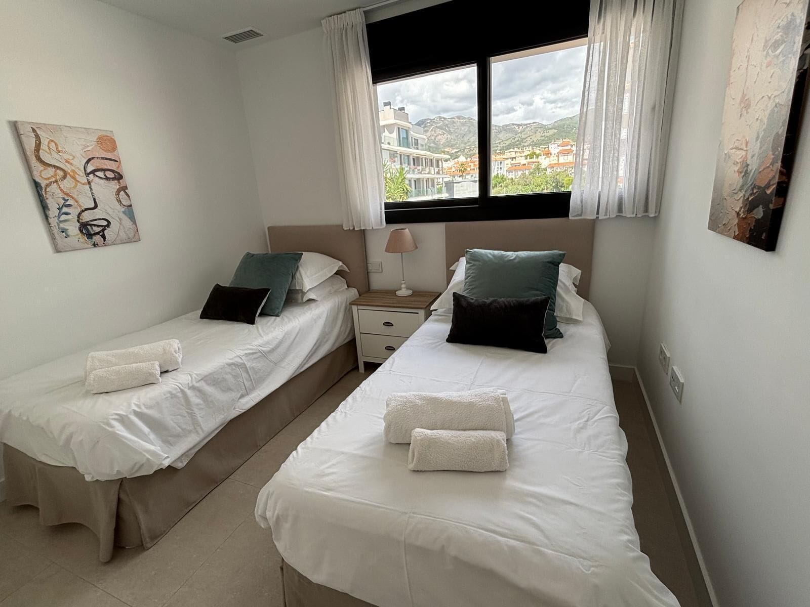 Ein Schlafzimmer mit zwei Betten, einem Nachttisch, einer Lampe und einem Fenster mit Blick auf Gebäude.