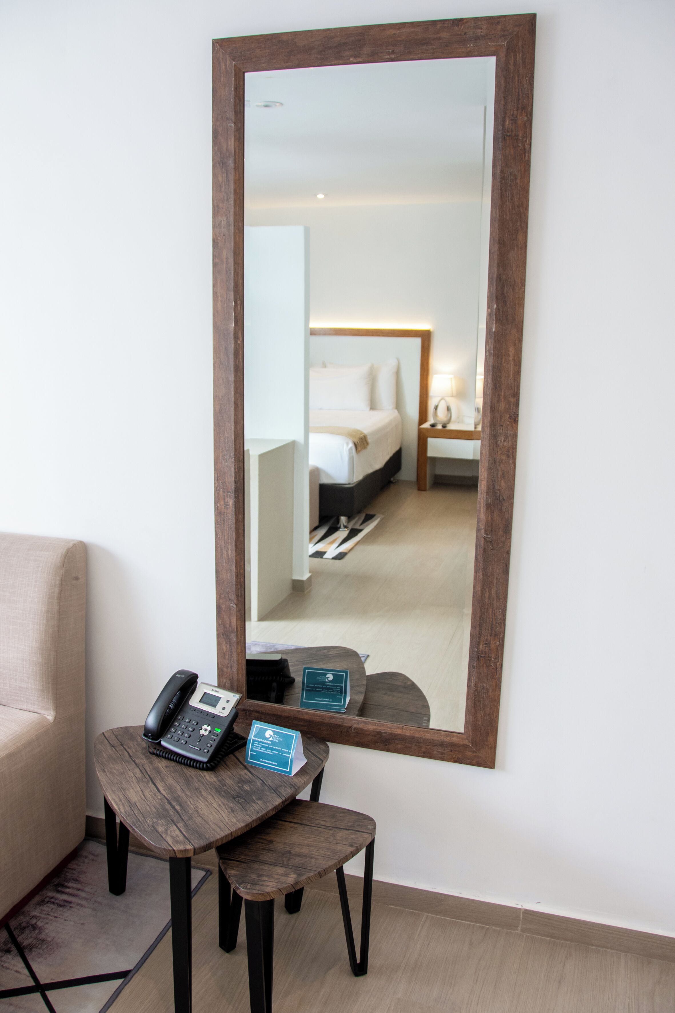Een hotelkamer met een grote spiegel, een nachtkastje met een telefoon en een bordje, een bed en een nachtkastje met een lamp.