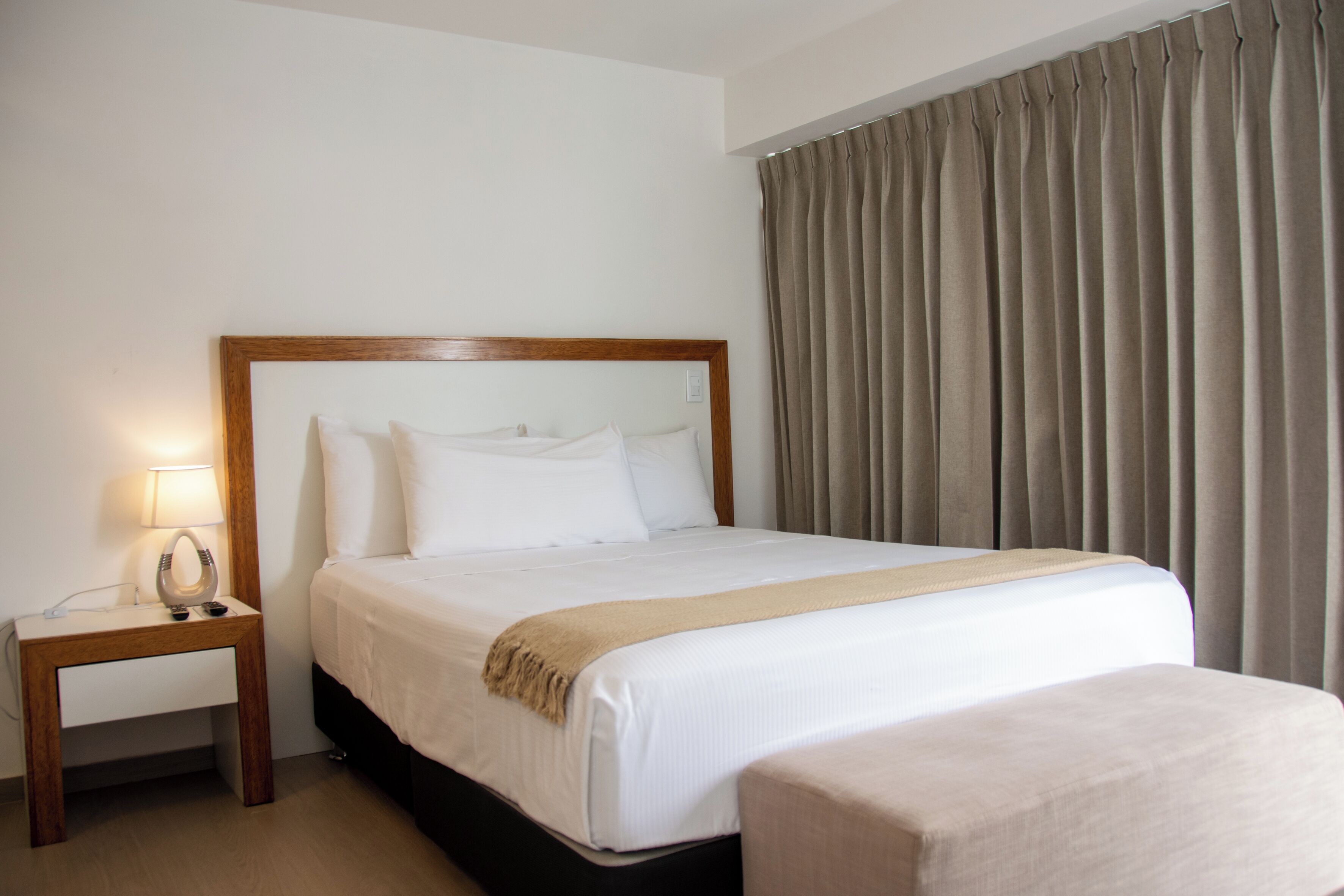 Una habitación de hotel con una cama grande, una mesita de noche con lámpara y una ventana con cortinas.