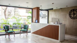 A hotel reception area with a modern design, featuring a reception desk, two green upholstered chairs, and a large wall clock. - GRAU BUSINESS HOTEL (Piura)