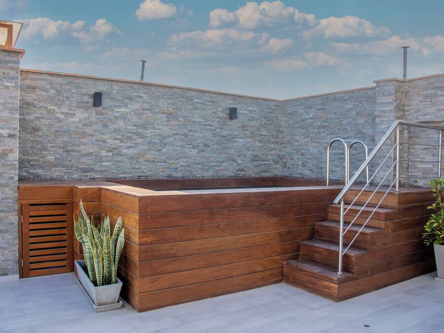 Una terraza en la azotea con un muro de piedra, un banco de madera, una planta en maceta y una escalera que lleva a un nivel superior.