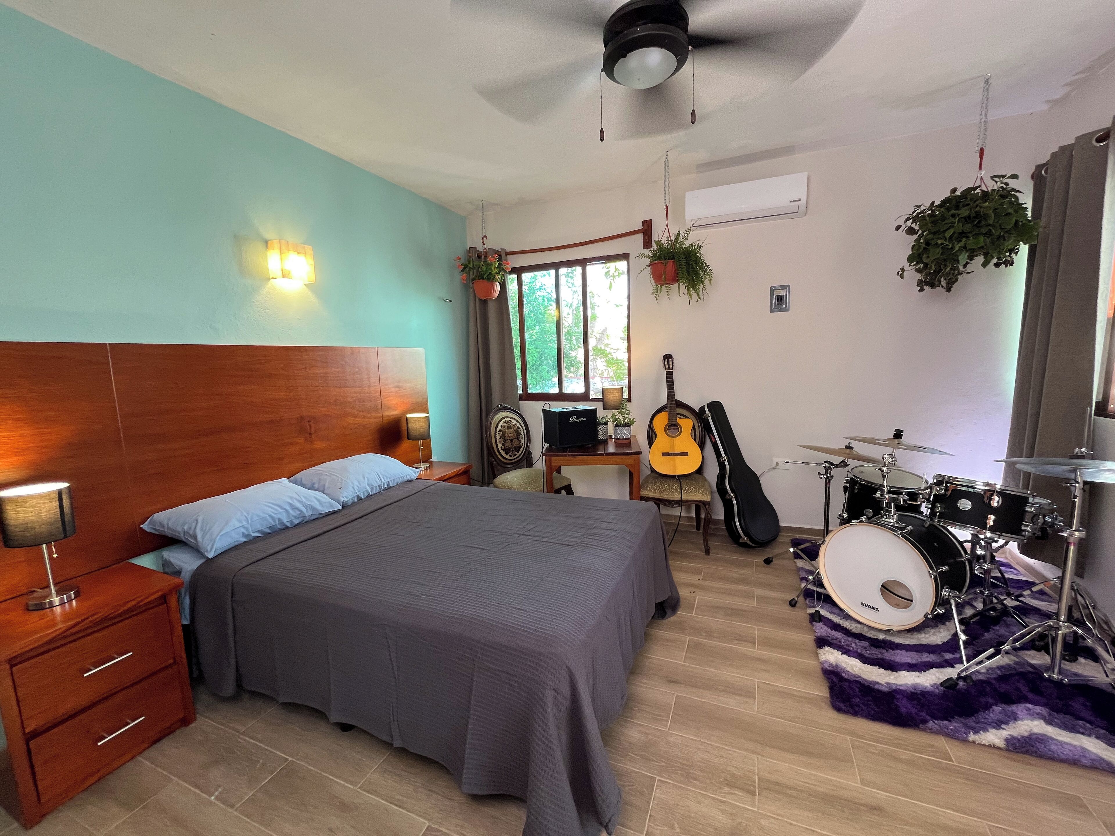 A bedroom with a bed, a desk, a guitar, a drum set, and a ceiling fan.
