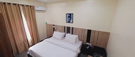 A hotel room with a bed, a nightstand, an air conditioning unit, and a framed picture on the wall.
