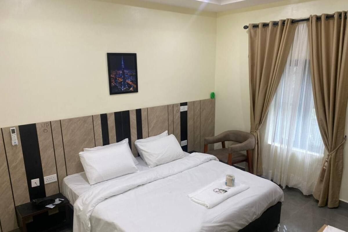 A hotel room with a bed, a chair, a nightstand, a picture on the wall, and a window with curtains.