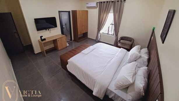 A hotel room with a bed, a desk, a chair, a TV, an air conditioning unit, and a window with curtains. - Victia Suite and Lounge (Ijebu Ode)