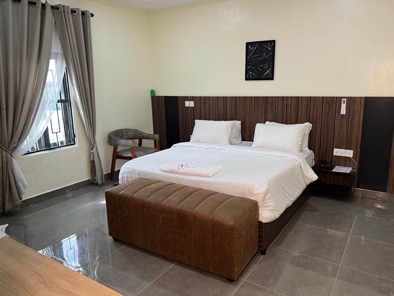 A hotel room with a large bed, a wooden headboard, a bedside table, a nightstand, a bench, and a window with curtains.