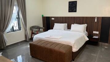 A hotel room with a large bed, a wooden headboard, a bedside table, a nightstand, a bench, and a window with curtains.