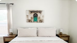 Une chambre à coucher comprenant un lit, deux tables de chevet, une commode, une fenêtre avec des rideaux et un tableau représentant un bâtiment, accroché au mur.