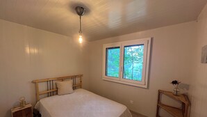 A bedroom with a wooden bed, a nightstand, a bedside table, a window with a view of trees, and a ceiling light. - Cocooning cottage in the heart of nature (Saxon)