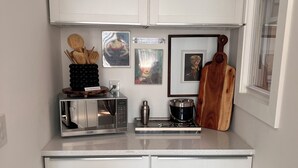 Una cocina compacta con armarios blancos, un microondas de acero inoxidable, una cafetera y una tabla de cortar de madera.