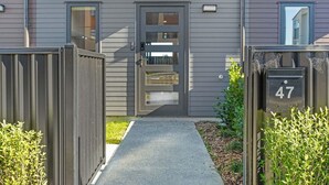 A modern building with a grey facade, a front door with a glass panel, and a small garden area with bushes.