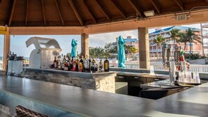 Zona de bar techada con una barra larga, taburetes y vistas a la playa y edificios al fondo.