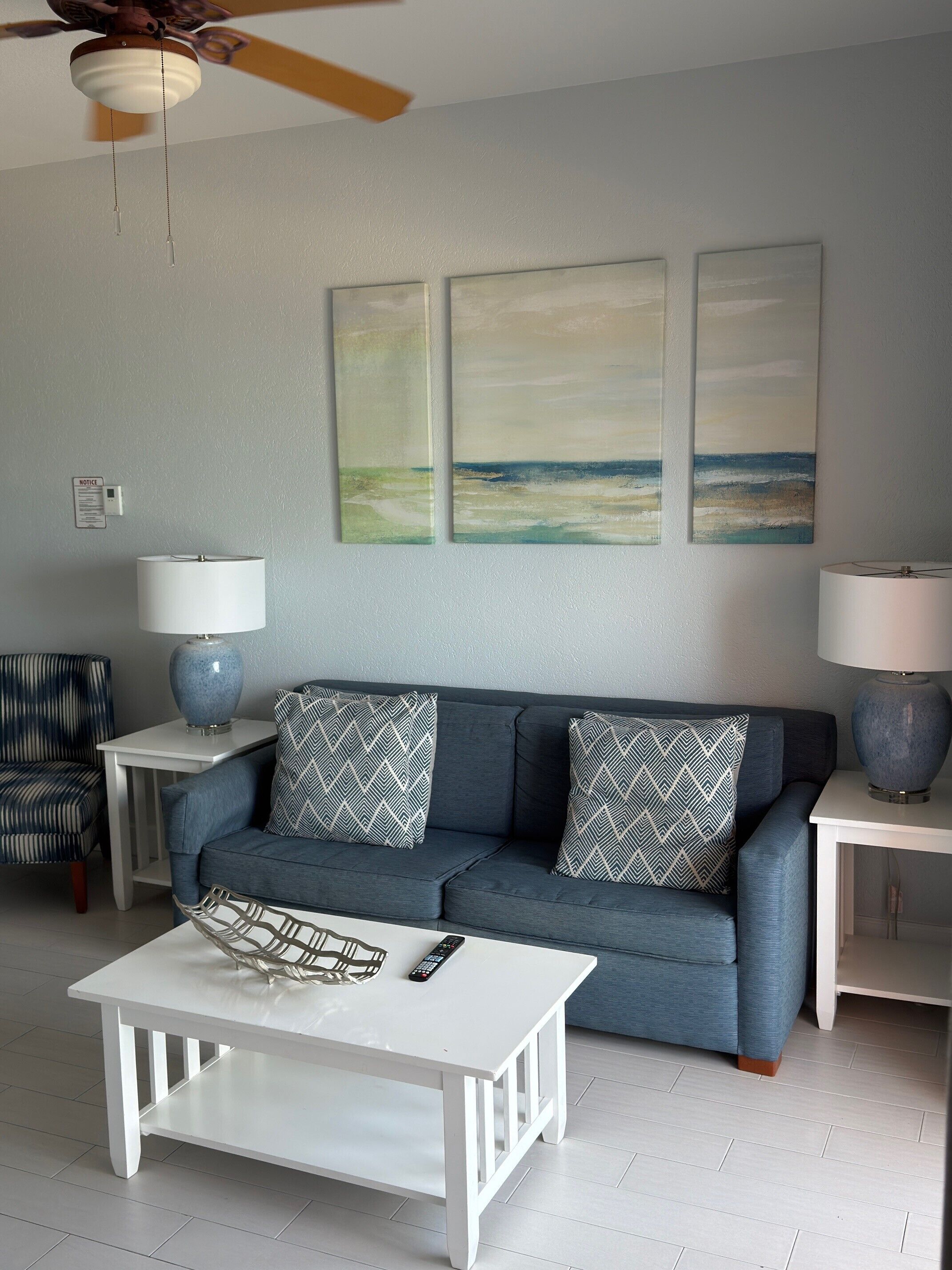 Una sala de estar con sofá, mesa de centro, lámpara, sillón y una obra de arte en la pared.